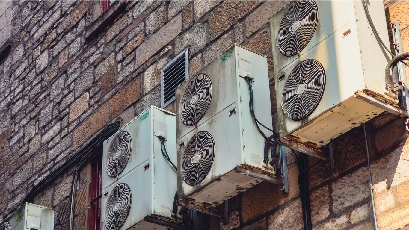 Multiple old, rusted air conditioning condenser units mounted on a wall, illustrating the need for A/C replacement due to age and inefficiency.