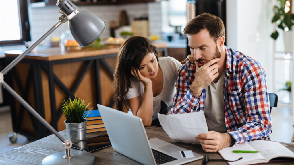 A stressed couple looking at a high energy bill in their kitchen, wondering why their electricity costs have spiked due to A/C issues.