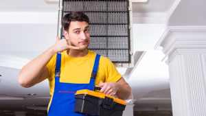An A/C repair technician in uniform holding a toolbox and making a "call me" gesture, with an air conditioning unit in the background.