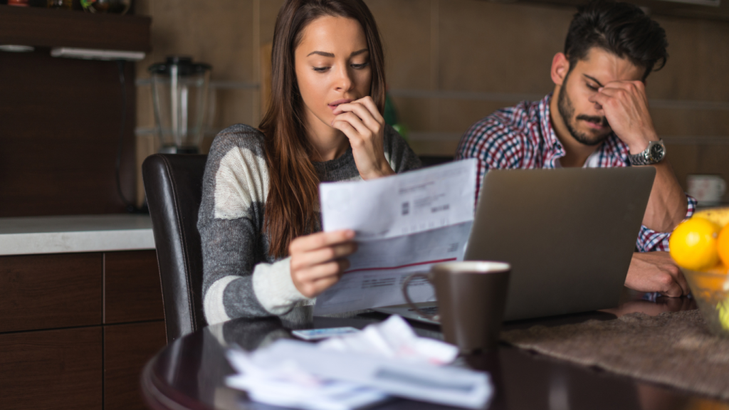 A young couple looking stressed and frustrated while reviewing their high electric bills at a kitchen table with a laptop.
