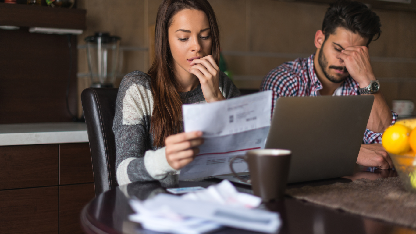 A young couple looking stressed and frustrated while reviewing their high electric bills at a kitchen table with a laptop.