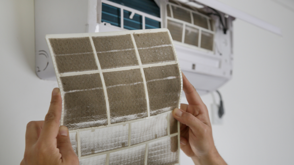 A person holding up a very dirty, clogged A/C filter in front of an open air conditioner unit, showing the cause of poor indoor air quality.