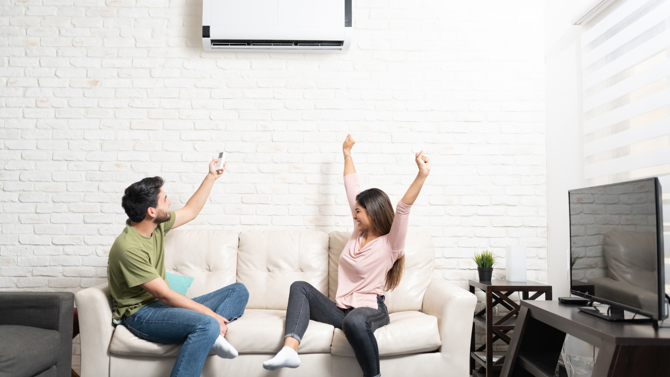A happy couple celebrating on their couch, using the remote to control their new wall-mounted ductless mini-split A/C unit, enjoying zoned comfort.