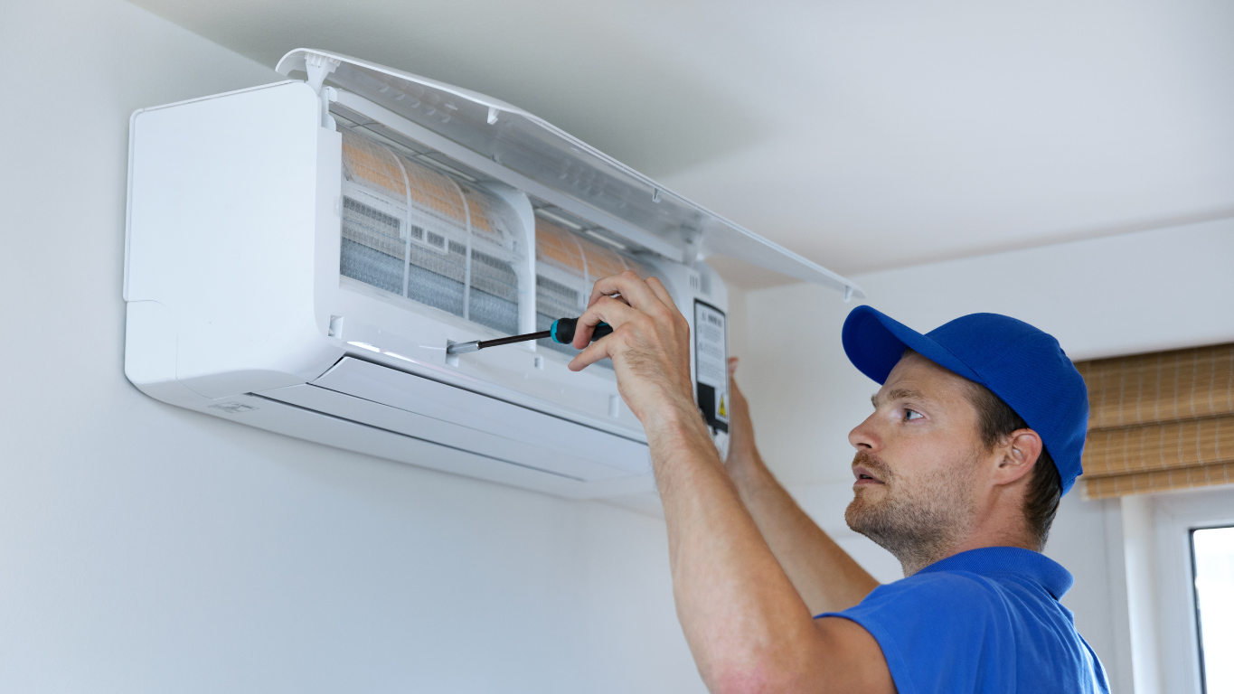 A professional HVAC technician in a blue cap installing or repairing an indoor, wall-mounted ductless mini-split A/C unit with a screwdriver.